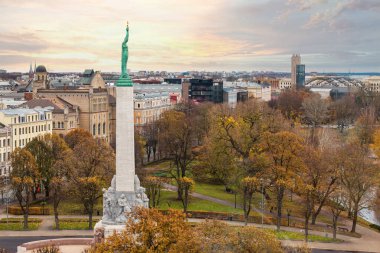 Letonya 'nın Riga kentindeki Özgürlük Anıtı sonbahar ağaçlarıyla çevrilidir. Şehir manzarası, pastel bir gökyüzü altında tarihi ve modern binaları harmanlıyor..