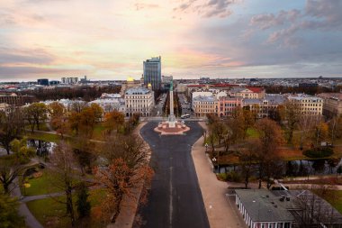 Riga, Letonya 'nın Özgürlük Anıtı, sonbahar ağaçlarıyla çevrili bir hava manzarası etrafında yer alıyor. Modern bir bina, pembe turuncu gökyüzü altında geleneksel mimariyle çelişiyor..