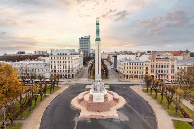 Özgürlük Anıtı Riga, Letonya 'da tarihi binalarla çevrili merkezi bir meydanda duruyor. Sonbahar ağaçları ve pastel bir gökyüzü sakin bir manzara yaratır..