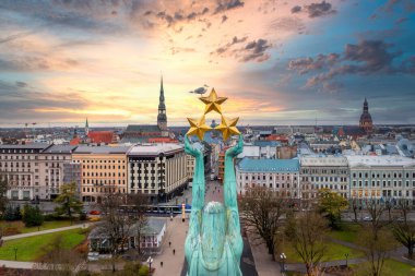 The Freedom Monument in Riga, Latvia, with a seagull perched on top, stands in the foreground. St. Peter's Church spire and city skyline are visible at sunset. clipart