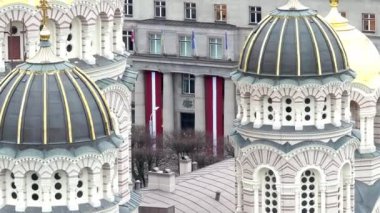 Resim, Letonya 'nın Riga kentindeki İsa' nın Doğumu Katedrali 'nin altın aksanlı süslü kubbelerini gösteriyor. Arka planda kırmızı ve beyaz pankartlar görünür..
