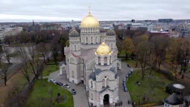 Riga, Letonya 'daki İsa' nın Doğuşu Katedrali 'nde bulutlu bir gökyüzü altında bir park ve şehir manzarasıyla çevrili altın kubbeler ve karmaşık mimari yer almaktadır..