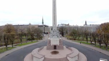 Özgürlük Anıtı, Letonya 'nın Riga şehrinde dairesel bir plazayla çevrili duruyor. Tarihi binalar ve bir kilise kulesi görülebilir, ağaçların oluşturduğu yollarla çerçevelenmiş..