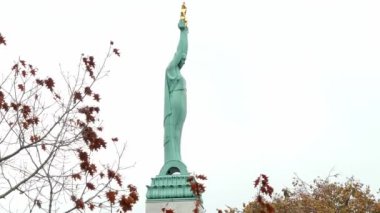 Riga, Letonya 'daki Özgürlük Anıtı üç yıldızlı Özgürlük Anıtı. Kahverengi yapraklı sonbahar ağaçları bulutlu bir gökyüzünün altında anıtın etrafını sarıyor..