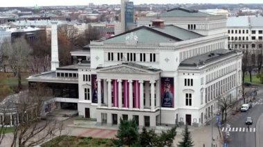 Riga 'daki Letonya Ulusal Operasının neoklasik cephesinde ağaçlar ve şehir sokaklarıyla çevrili uzun kolonlar ve afişler yer alıyor..