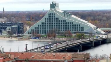 Riga 'nın şehir manzarasında modern Ulusal Kütüphane, Aziz Peter Kilisesi kulesi ve sonbahar ağaçları tarafından çerçevelenmiş bir köprüsü olan Daugava Nehri yer alır..