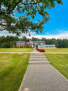 Gösterişli tasarımı olan çağdaş bir ev, otlarla çevrili asfalt bir yolun sonunda yer alır. Arkasında ağaçlar olan bir ağaç dalı tepeyi çerçeveler..