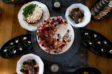 Çilek, yabanmersini ve çiçeklerle süslenmiş, çeşitli aperatifler ve ahşap bir yüzeyde yanmamış mumlarla çevrili dekoratif bir pasta seti..