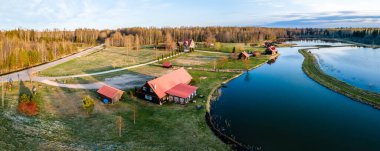 Bir grup kırmızı çatılı bina, açık tarlalarla çevrili dolambaçlı bir su yolunun yanında ve berrak bir gökyüzünün altında yapraksız ağaçlardan oluşan sık bir ormanın içinde..