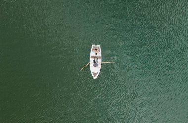 Yukarıdan bakıldığında yeşil suda küçük bir kayık görünüyor. Biri kürek çekerken diğeri hafif dalgalarla çevrili ve yalnızlığı vurguluyor..