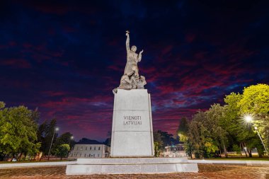 The Vienoti Latvijai monument stands in a lit square in Rezekne, Latvia, with a figure holding a star against a red and purple evening sky. clipart