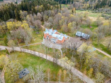 An aerial perspective captures a large brick mansion with symmetrical design, surrounded by deciduous and coniferous trees, open grassy areas, and nearby outbuildings. clipart