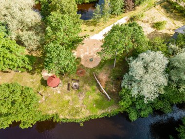 An aerial view captures a secluded riverside area with a red roofed gazebo, fire pit, and log seating, surrounded by lush trees and a gently curving river. clipart