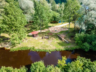 Bir parkta bir nehir, kırmızı çatılı bir çardak, piknik bankları ve bir oyun alanı bulunur. Ağaçlar gölgeyi sağlar, aile dostu bir ortam yaratır..