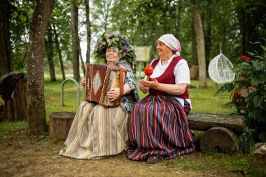 Halk kostümlü iki yaşlı kadın ormanda bir bankta oturuyor. Biri çiçekli bir taç ile akordeon çalarken, diğeri kırmızı bir maraca tutar..