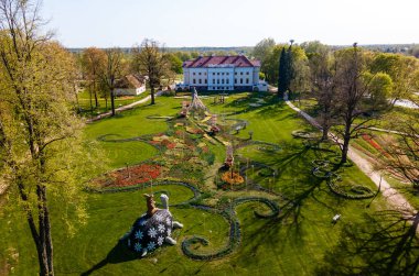 Pakruojis Malikanesi, beyaz cephesi ve kırmızı çatısı ile renkli lale yataklarıyla ve bir kaplumbağa da dahil olmak üzere tuhaf heykelleriyle çevrili..