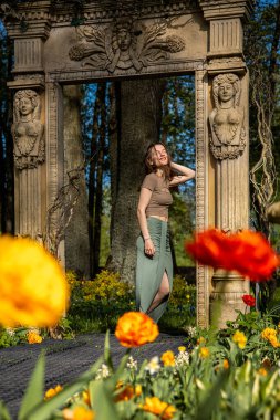 Taştan bir kemerin altında heykel figürleriyle bir kadın duruyor. Etrafı parlak sarı ve kırmızı lalelerle çevrili, uzun ağaçlara karşı dizilmiş..