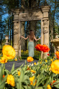 A woman stands in a garden of yellow and orange tulips, in front of a stone archway with intricate carvings, surrounded by tall trees. clipart