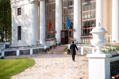 A grand neoclassical building features tall white columns and large windows. Flags in red and blue yellow are displayed. A formally dressed person walks towards the entrance, with a decorative urn and greenery lining the pathway.