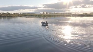 Bir insan geniş bir gölde kürek çeker. Güneş ışığı gökyüzünde bulutlarla birlikte suyu yansıtır. Ufuk çizgisinde ağaçlar ve yakınlarda bir şamandıra yüzüyor..