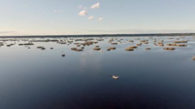 Yalnız bir tekne, geniş bir su boyunca hareket eder. Küçük adalar ve kamışlarla birlikte. Dağınık bulutlu açık gökyüzü sudaki sakin bir günü işaret ediyor..