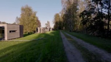 Minimalist mimarisi ve geniş pencereli yazlık evler çimenlik bir yol çiziyor. Bisikletler dışarıda park halinde ve uzun ağaçlar uzun gölgeler oluşturuyor..