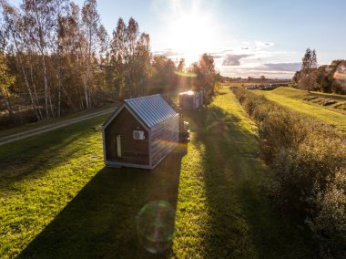 Metal çatılı küçük, modern bir kulübe çimenli bir alanda yer alır. Ağaç ağaçları sol tarafta, ufuk çizgisine uzanan açık alanlar sıcak bir parıltının altında..