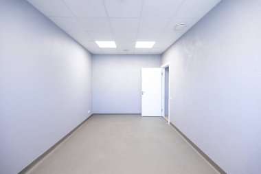 Minimalist bir odada açık gri duvarlar ve uygun bir zemin bulunur. Işıklandırma panelleri boşluğu aydınlatıyor. Sağda beyaz bir kapı var..