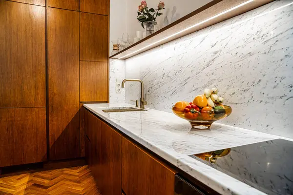 Modern bir mutfakta zengin ahşap dolaplar, mermer tezgah ve pirinçten bir musluk bulunur. Bir kase meyve ve içinde güller olan bir vazo renk ve zarafet ekler..