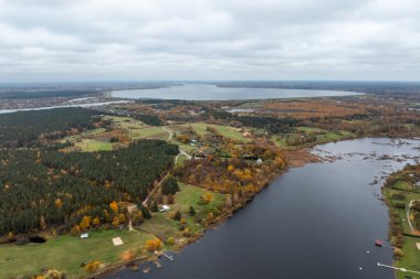 Geniş bir hava manzarası, canlı turuncu ve sarı ağaçlarla çevrili sonbahar yapraklarıyla çevrili büyük bir göl gösteriyor. Yollar ve küçük binalar kırsal bir ortamı gösteriyor..
