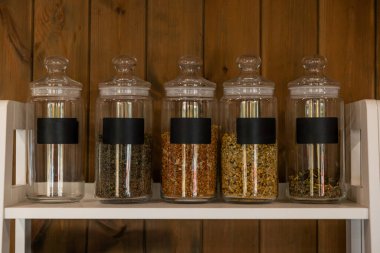 Siyah etiketli beş adet cam kavanoz, beyaz bir rafta duran çeşitli kurutulmuş otlar içeriyor. Ahşap duvar düzenlemeye kırsal bir unsur ekler..