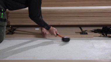 Kapalı alanda ahşap döşeme tahtalarını güvence altına almak için lastik çekiç kullanan bir insan, arka planda kalas yığınları ile işçiliği ön plana çıkarıyor..