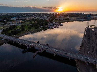 Güneş Letonya, Ventspils üzerinde batıyor, nehir ve köprüye sıcak bir parıltı saçıyor. Araçlar köprüyü geçiyor, ufukta Baltık Denizi görünüyor..