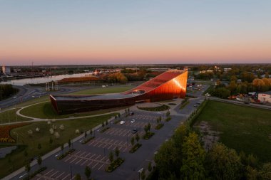 Letonya 'daki Ventspils Bilim ve İnovasyon Merkezi bakır cepheli modern bir kavisli tasarıma sahiptir. Arka planda bir nehir ve şehir manzarası görülebilir.