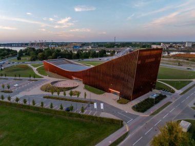 Ventspils Konser Salonu Latvija açısal tasarımını ve pas cephesini sergiliyor, yeşil alanlar ve yollar arasında ayarlanıyor, vinçler uzaktan görülebiliyor..