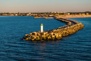 Beyaz bir deniz feneri ve yeşil kapaklı uzun bir dalgakıran denize doğru uzanıyor. Kumlu plajı ve arka planında binaları olan yapıyı köşeli beton bloklar oluşturur..