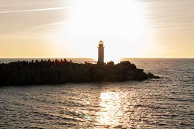 Batan güneşin siluetiyle kayalık bir dalgakıranın üzerinde bir deniz feneri duruyor. Güneşin yansıması, üzerinde açık bir gökyüzü olan, suyun üzerinde parıldayan bir yol yaratır..