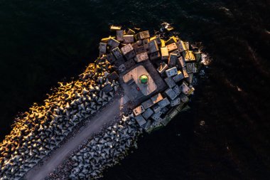 An aerial perspective of a breakwater featuring interlocking concrete blocks and a green lighthouse, set against deep, dynamic waters under warm sunlight. clipart