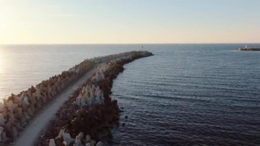 Beton tetrapodlu uzun bir dalgakıran, içinde küçük bir deniz feneri bulunan denize doğru uzanır. Uzak bir dalgakıranda başka bir deniz feneri görünür..