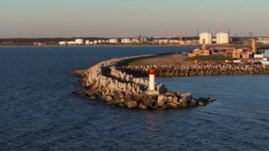 Beton tetrapodları ve kırmızı tepeli deniz feneri olan bir dalgakıran, arka planda endüstriyel tanklar ve yapılarla sakin sulara doğru uzanır..