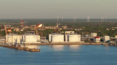 Letonya, Ventspils 'te bir liman yakınında' VENT 'etiketli büyük beyaz depolama tankları. Turnalar, rüzgar türbinleri ve iletişim kulesi endüstriyel elementleri vurguluyor.