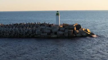 Yeşil tepeli bir deniz feneri, sıcak bir gün batımının altında sakin deniz sularıyla çevrili geometrik beton bloklardan oluşan bir dalganın üzerinde duruyor..