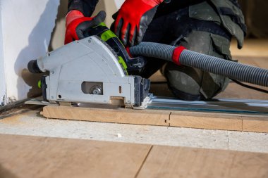 Kırmızı eldivenli bir insan, kapalı alanda tahta testereler üzerinde dairesel bir testere kullanır. Koruyucu teçhizat ve toz çıkarma hortumu marangozluk işinde güvenliği vurguluyor.