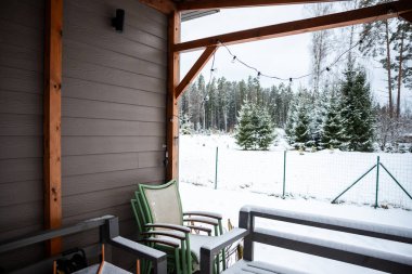 A terrace with wooden beams, LED lights, and snow covered furniture, set against a backdrop of evergreen trees and a snowy forest. clipart