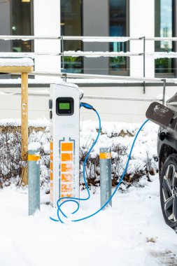 Kar kaplı bir istasyonda turuncu aksanlı, kalın balinalarla çevrili elektrikli bir araç şarj oluyor. Arka planda geniş pencereli modern bir bina var..