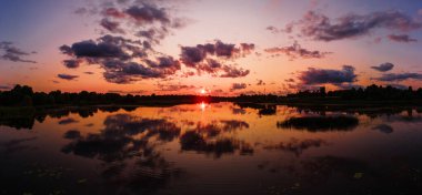 Gün batımında ufukta turuncu ve pembe renkler belirir. Dağılmış bulutlar ve su bitkileri simetrik yansımayı arttırır.