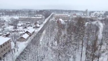 Düz bir yol, daha küçük evler, apartmanlar, bacası tüten bir fabrika ve karla kaplı bir çevreye sahip kışın kent manzarası..