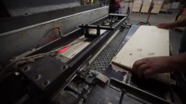 Bir el, loş ışıklı bir atölyede kırmızı lazer rehberi olan bir makineden hafif renkli ahşabı geçirir. Tahta sandıklar ve endüstriyel ekipmanlar görülebilir..