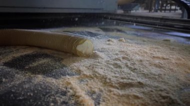 Süpürge, atölyedeki karanlık bir yüzeyden talaş temizler. Yakınlarda bulanık bir hortum var. Arka planda da bulanık makineler var..