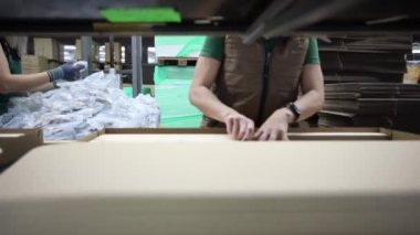 Bireyler karton kutuları birleştirir ve plastik ambalaj malzemelerini düzenli malzeme yığınları ve görünür raflarla bir depoda tutarlar..